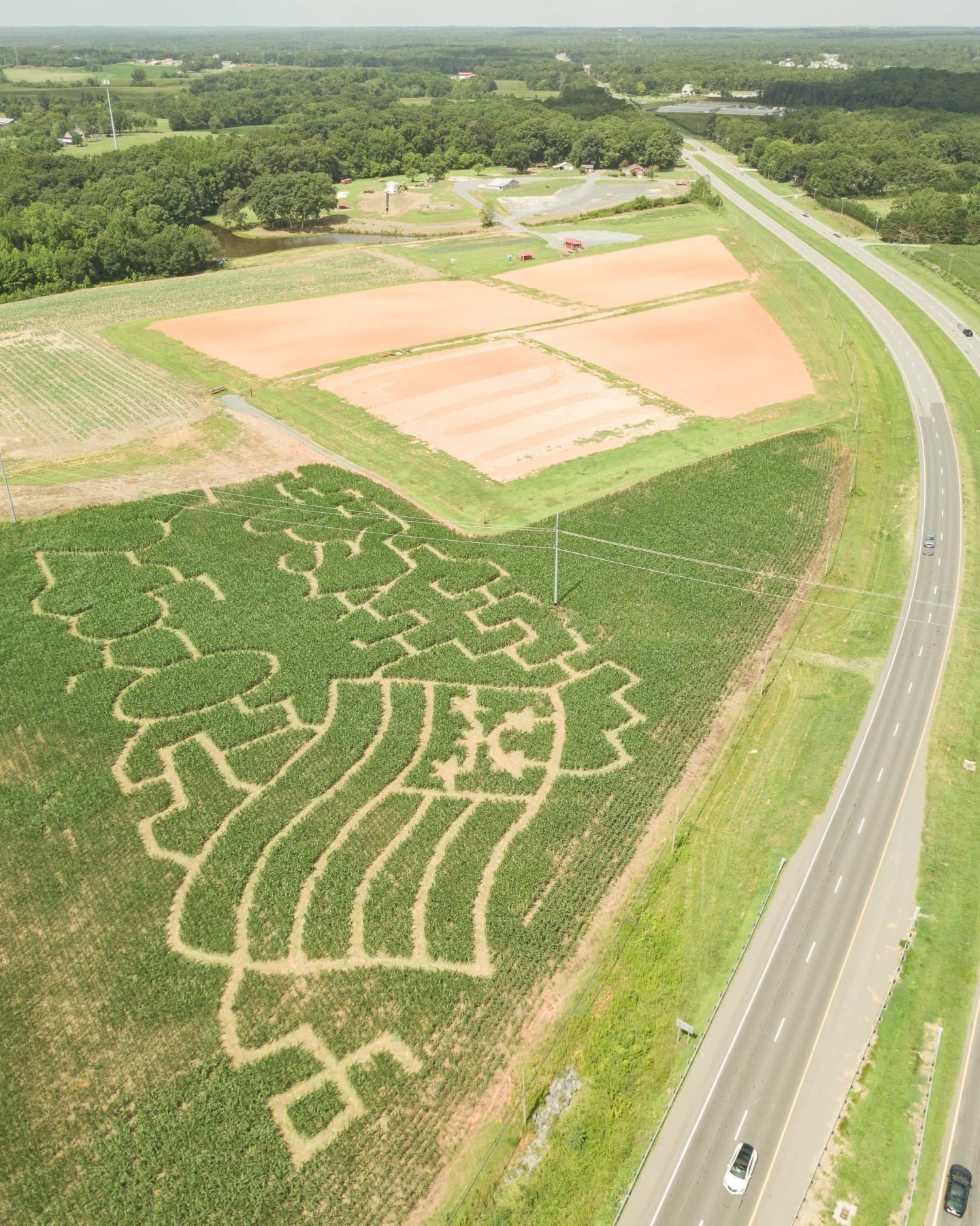 Wide aerial view of the Deer Run Farm corn maze beside the highway with fields, farm grounds, and wooded countryside beyond